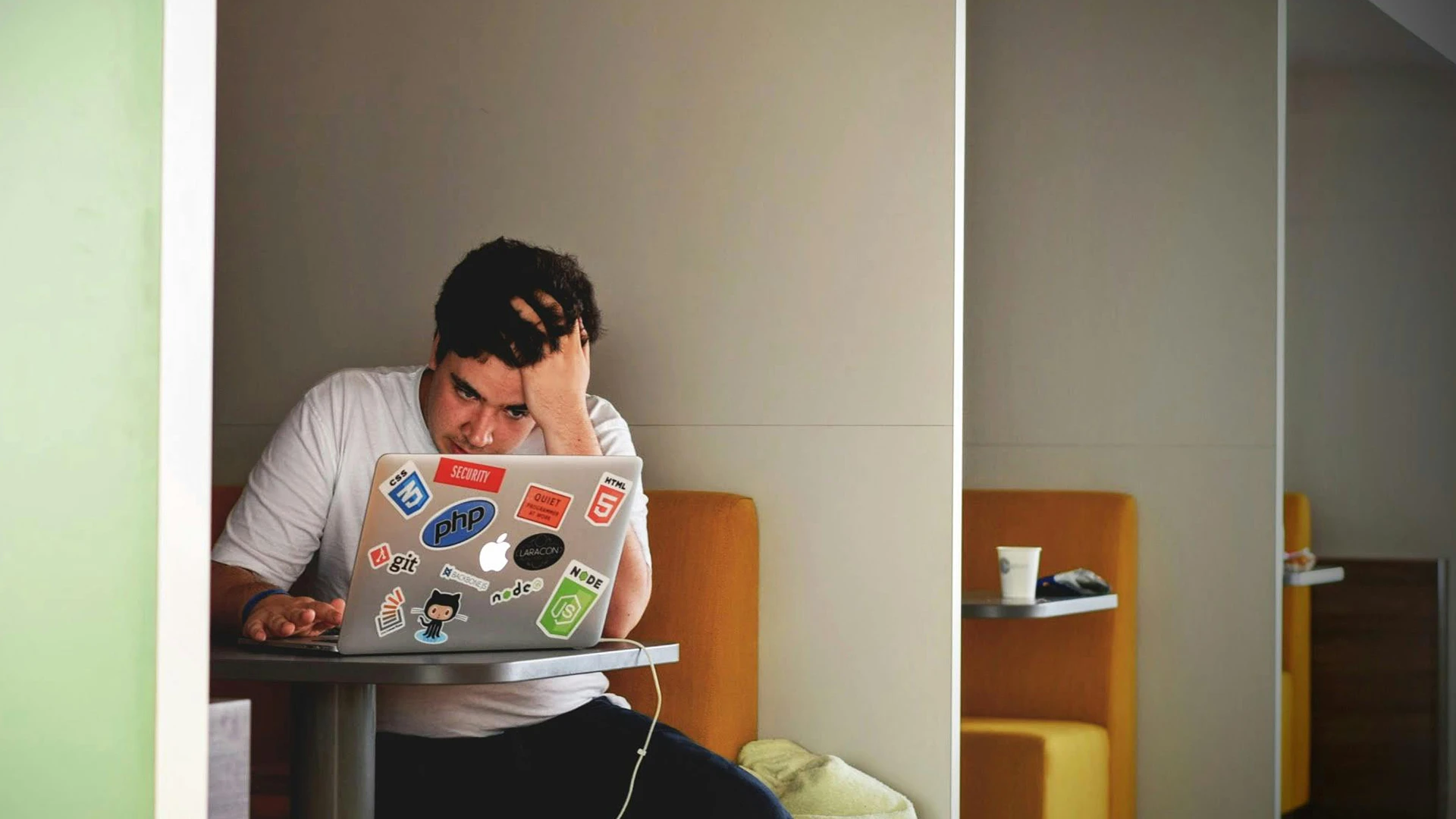 Frustrated man sitting at a table with his hand on his head, using a sticker-covered laptop in a modern seating area, symbolising challenges with slow website load times.